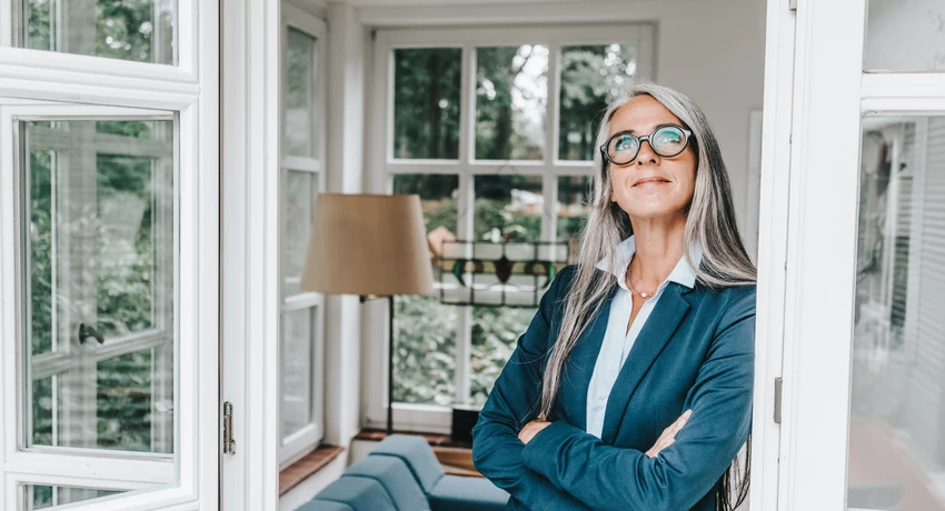 Woman in a blue blazer standing with arms crossed in a bright residential living space, framed by open glass doors and large windows.