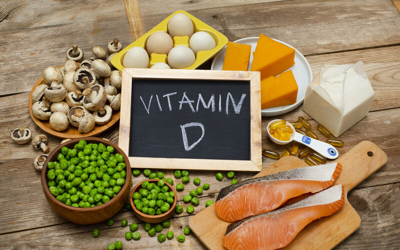 A variety of vitamin D-rich foods displayed on a wooden table, including mushrooms, eggs, cheese, milk, green peas, salmon fillets, and vitamin D supplements. A small chalkboard in the center reads 'Vitamin D.