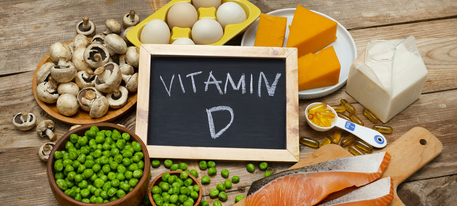 A variety of vitamin D-rich foods displayed on a wooden table, including mushrooms, eggs, cheese, milk, green peas, salmon fillets, and vitamin D supplements. A small chalkboard in the center reads 'Vitamin D.