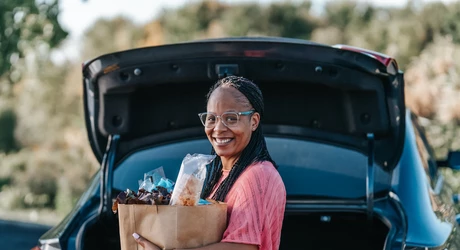 Person standing at the open trunk of a car holding a grocery bag, with trees and an outdoor setting in the background. 