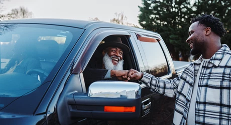 Senior man in blue truck fist bumps younger man who is standing outside truck.  