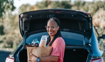 Person standing at the open trunk of a car holding a grocery bag, with trees and an outdoor setting in the background. 