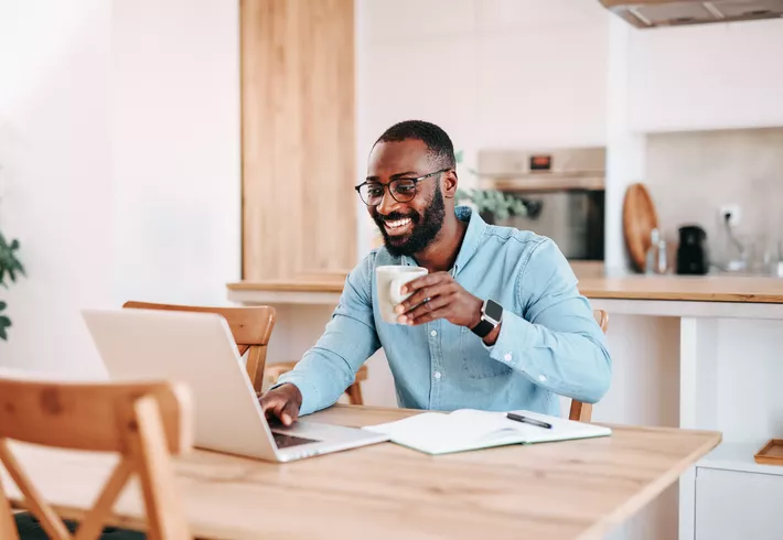 Person sitting at a table using a laptop and holding a mug, with a notebook and kitchen area in the background. 
