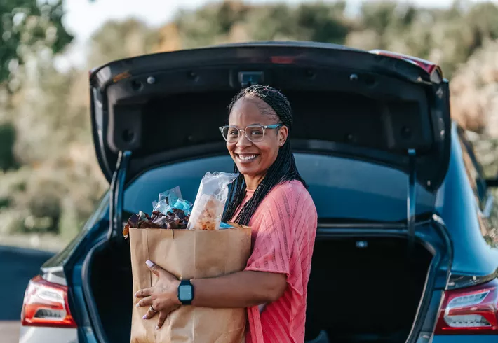 Person standing at the open trunk of a car holding a grocery bag, with trees and an outdoor setting in the background. 
