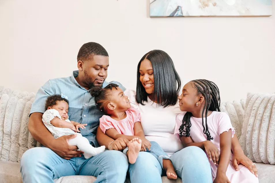A family seated on a couch, including an infant being held and two children sitting between their parents.
