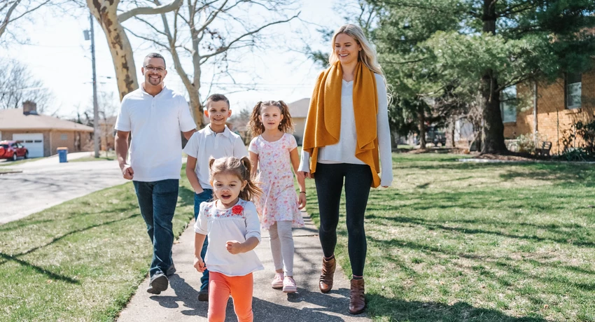 Five people walking together on a sunny suburban sidewalk, including two adults and three children. A small child runs ahead in the foreground, while the others walk behind along a tree-lined street with houses and grass lawns. 