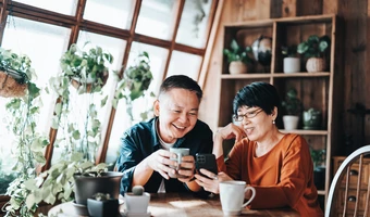 Two people sitting at a wooden table in a plant-filled home, looking at a smartphone together while enjoying hot drinks.