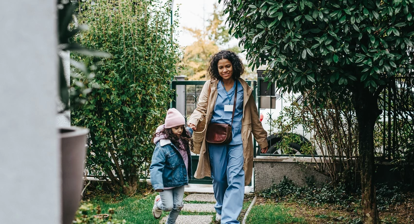 African American nurse in scrubs walking in yard, holding hands with her young daughter 