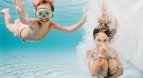 Two children underwater in a swimming pool, one wearing swim goggles. 