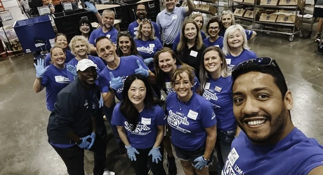 Group of volunteers in blue shirts gathered in a warehouse fulfillment center, posing for a photo among shelves of packaged goods and donation supplies.