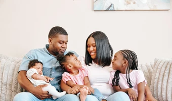 A family seated on a couch, including an infant being held and two children sitting between their parents.