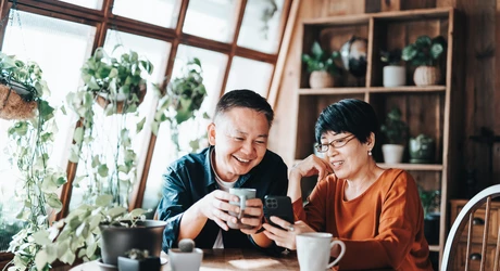 Two people sitting at a wooden table in a plant-filled home, looking at a smartphone together while enjoying hot drinks.