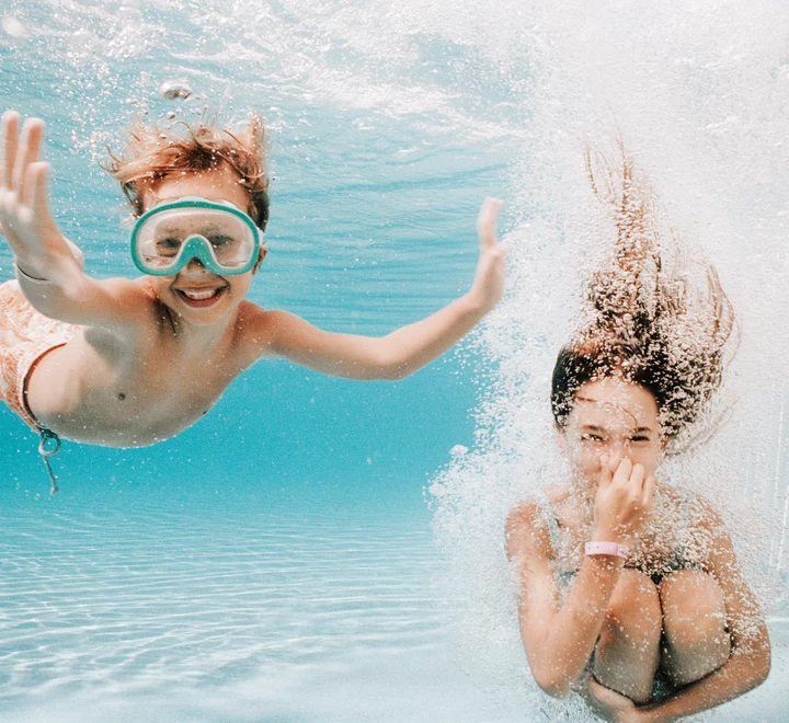 Two children underwater in a swimming pool, one wearing swim goggles. 