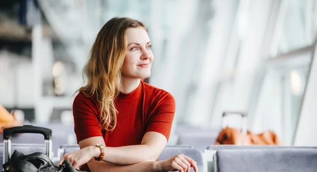 woman with strawberry-blonde hair smiles while sitting next to her luggage, looking out the window and waiting for her flight in an airport lounge. 