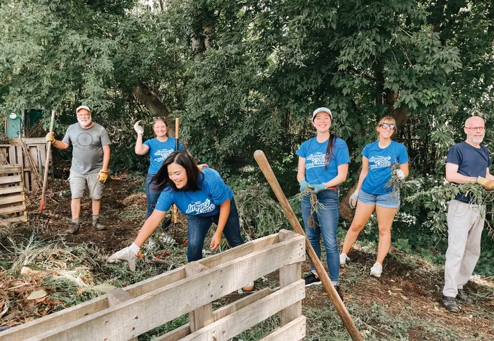 Group of volunteers working outdoors in a wooded area, clearing brush and debris near a wooden fence. 