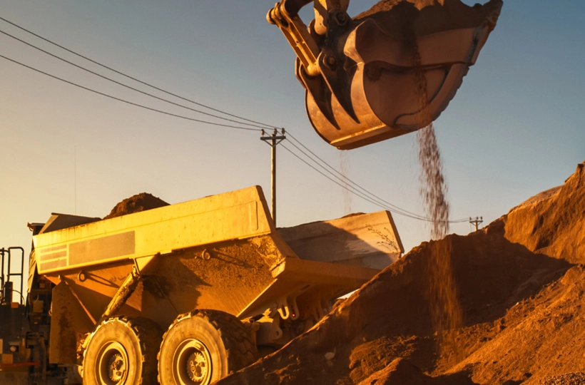 A large construction vehicle, specifically a dump truck, being loaded with dirt or sand by an excavator during sunrise or sunset. The warm lighting and long shadows create a dramatic effect. Power lines and poles in the background suggest that this activity is taking place near an urban area or infrastructure development site.