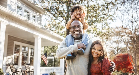 Father with two children standing in front of a home. The youngest child is on top of the father’s shoulders. The older child is standing next to him; father has his arm around the child. Everyone is smiling.  