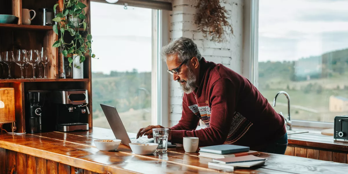 Older man on his laptop at a wooden kitchen counter. He is wearing a burgundy sweater.  