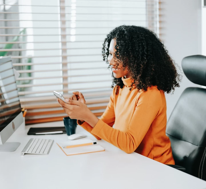Professional African American woman sitting at desk in office looking at cell phone 