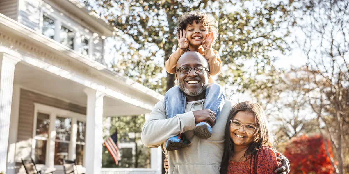 Father with two children standing in front of a home. The youngest child is on top of the father’s shoulders. The older child is standing next to him; father has his arm around the child. Everyone is smiling.  