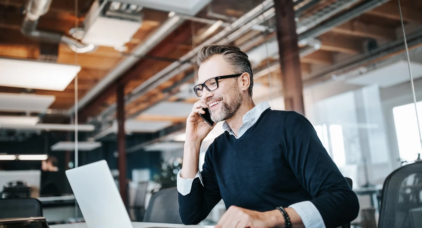 Smiling professional man sitting at desk on phone in windowed office    