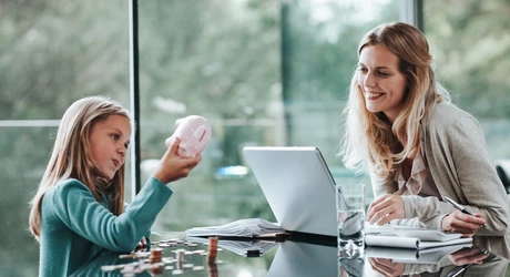Child holding a piggy bank with coins on the table while another person uses a laptop nearby. 