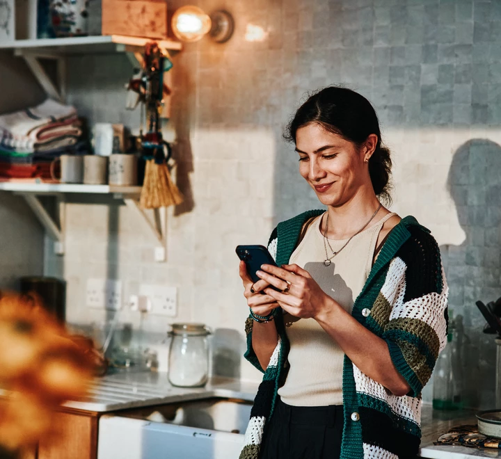 Smiling woman in her 30's standing in kitchen on smart phone. 