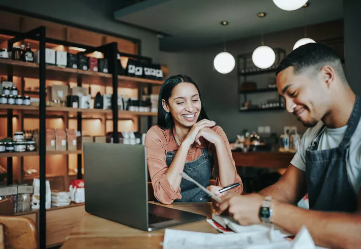 Two business owners going over paperwork and working on a laptop at a table in their shop    
