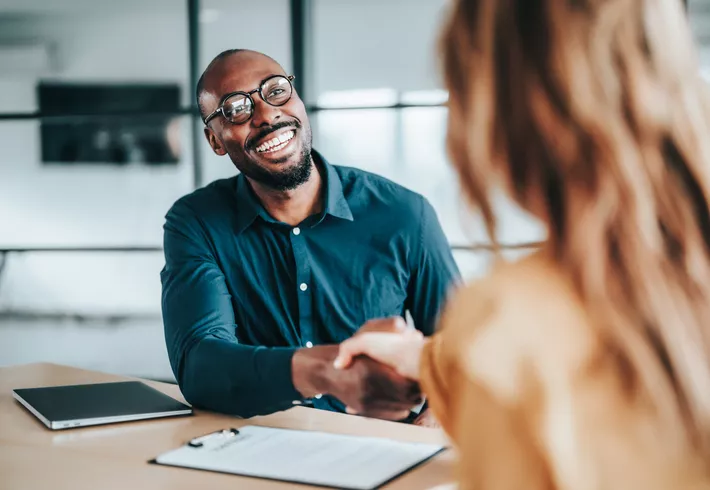 Two people shaking hands across a desk during an office meeting.
