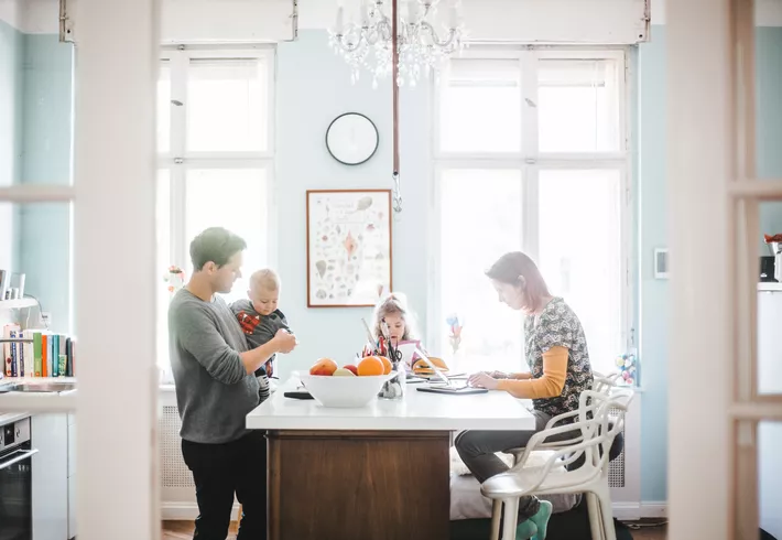 Bright kitchen with large windows. A person holds a toddler at the counter while another child draws and another person works on a laptop. 