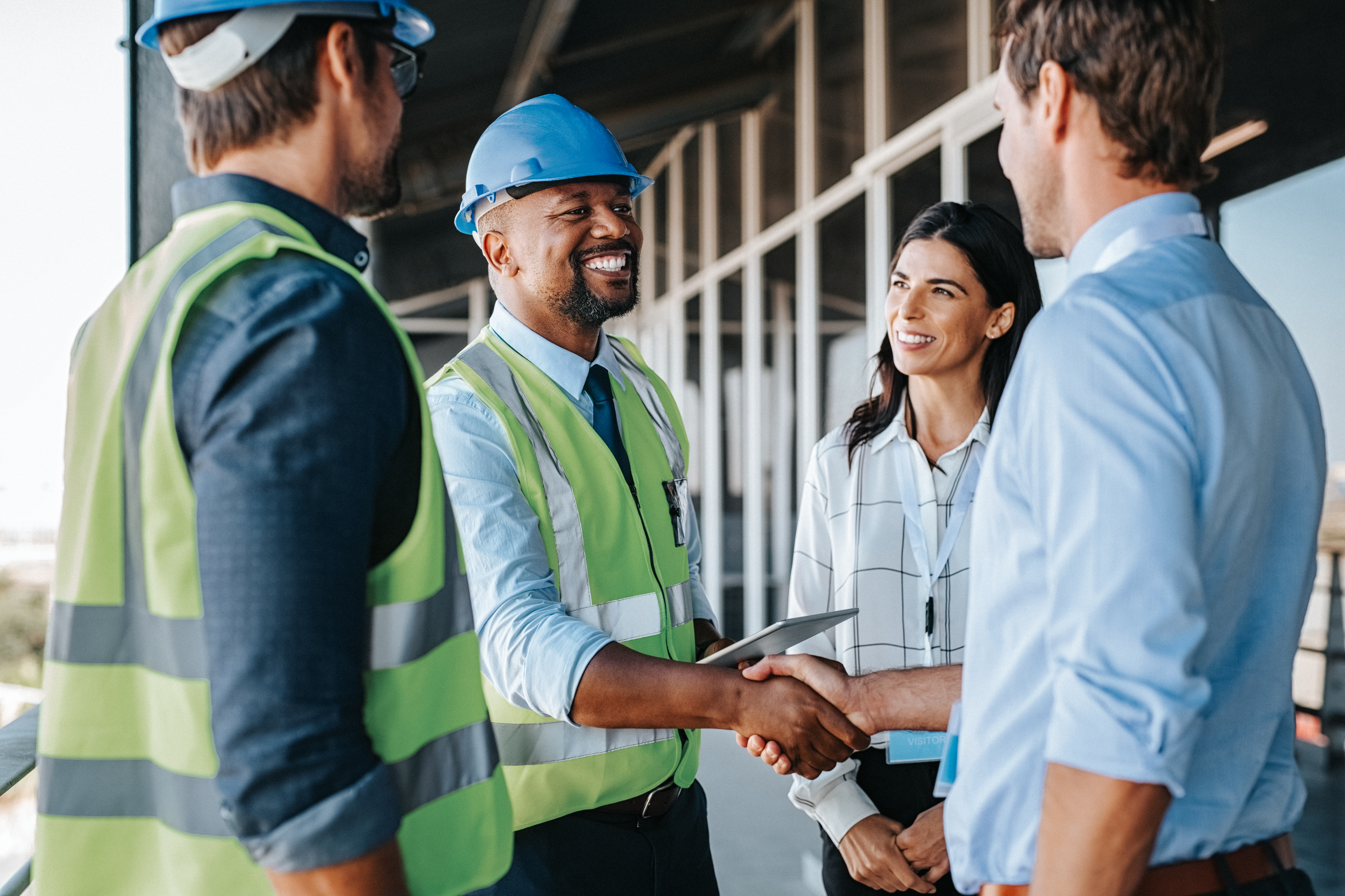 Smiling African American business man in hardhat and high visibility vest shaking business man's hand standing outside of building construction. 