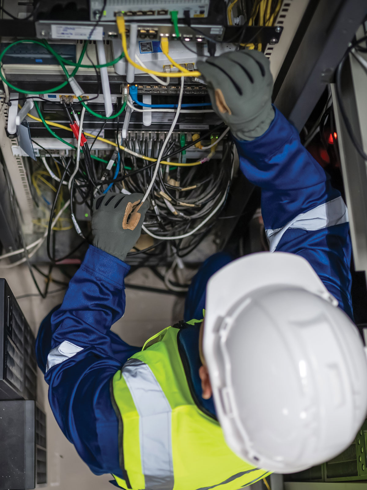Network technician in a hard hat and high-visibility vest patching Ethernet cables into a server rack