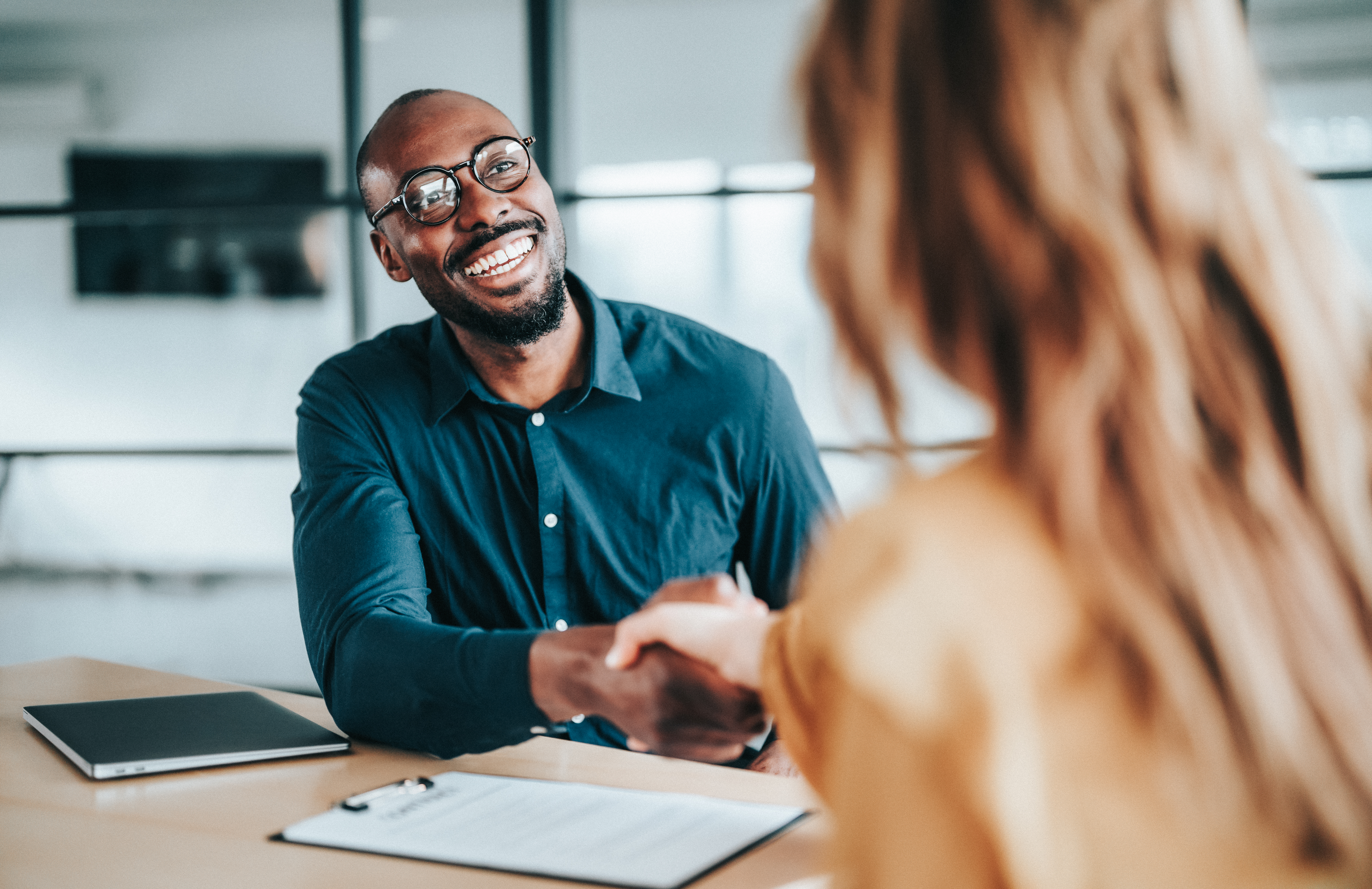 Two people shaking hands across a desk during an office meeting.