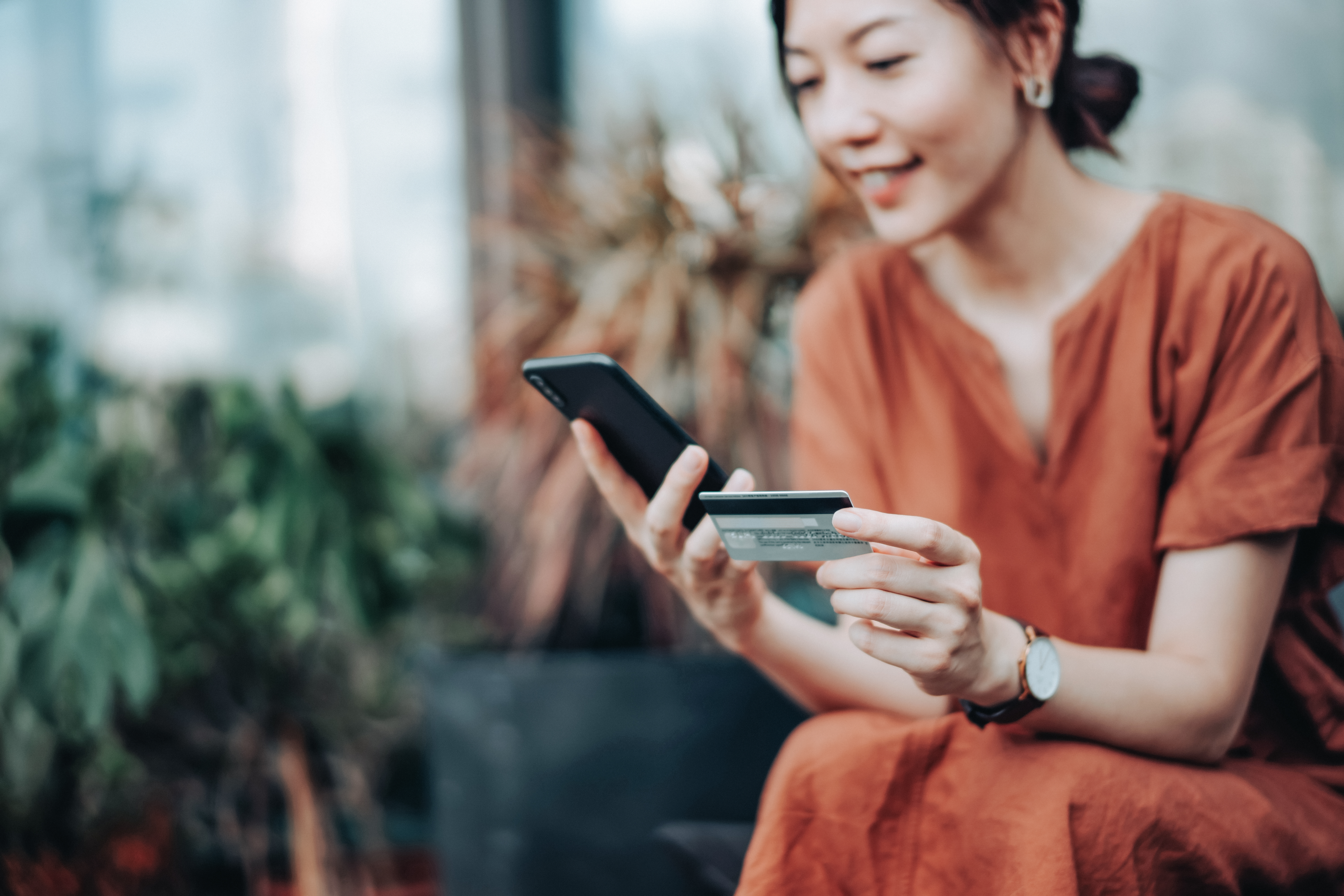 Young Asian woman sitting outside looking at cell phone and entering credit card information 