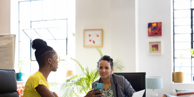 Two female workers sharing images on a smart phone in an office