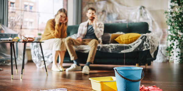 A white man and woman sit on a couch looking stressed while water leaks from their ceiling into a bucket on the floor