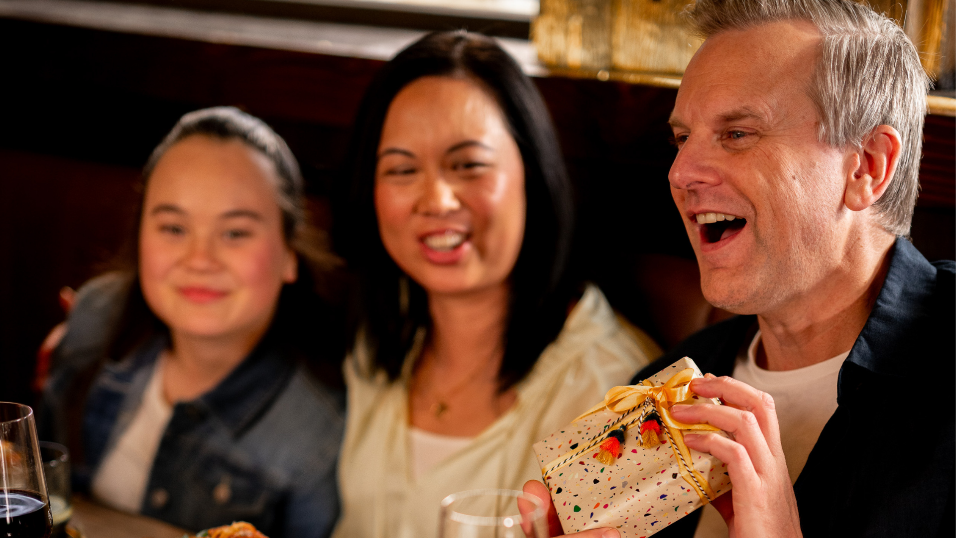 Dad holding a wrapped gift during a Father’s Day family meal at a restaurant with his wife and daughter.