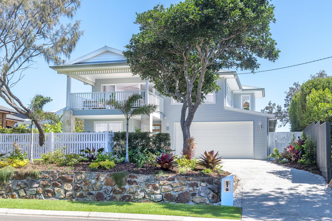 External view of the front of the house from directly in front. The house is two stories, with a large white garage on the right side, front porch on the left side. On the upper level a large covered entertainment deck is visible. There is a driveway on the right. A stone garden wall and gardens are in front of the white wooden fence. A large tree is in the front garden.
