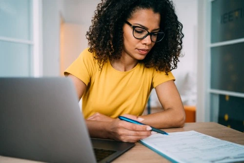 Woman in a yellow shirt writing on a form while working at a laptop in a bright, modern office.