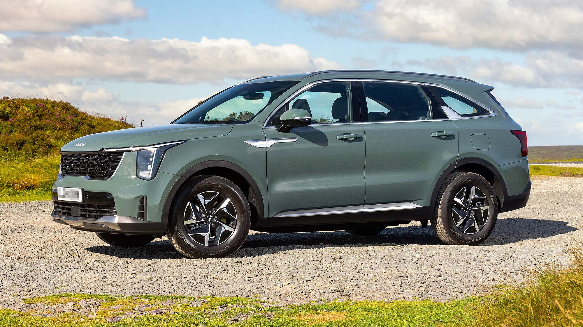 A green Kia Sorento SUV parked on a gravel surface in a countryside location, featuring a bold grille and spacious 7-seater body style.