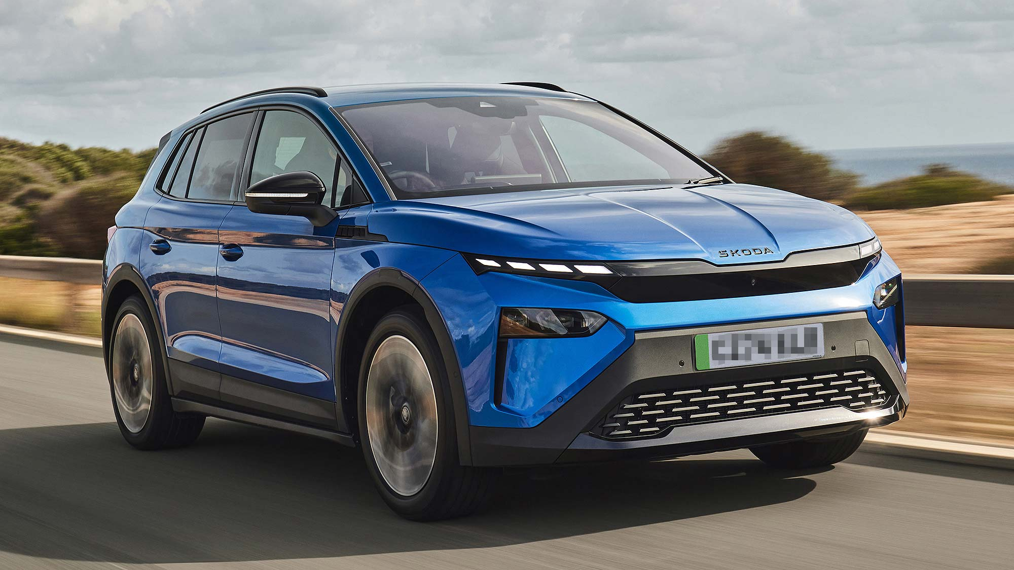 Blue Škoda Elroq electric SUV driving on a coastal road with blurred background, showcasing its sleek and modern design.