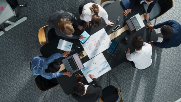 Top-down view of corporate team members standing around a conference table analyzing business metrics and site reports