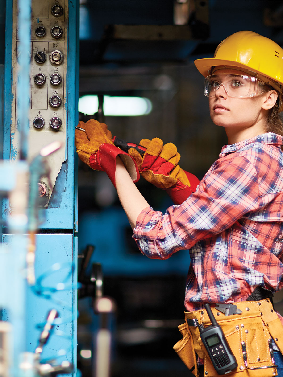 Female technician wearing a hard hat, safety glasses, and tool belt using a screwdriver on an industrial control panel