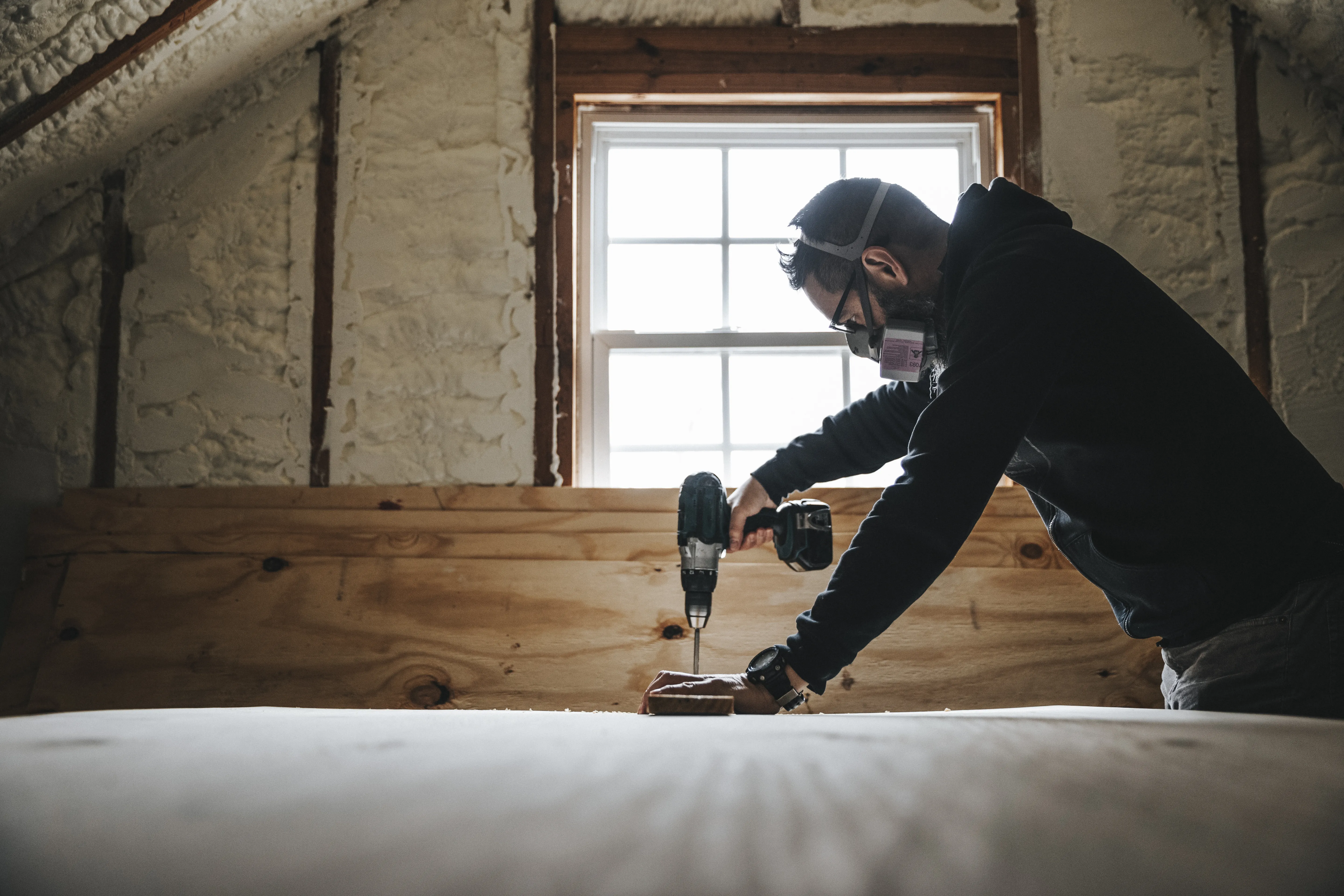 Man wearing a respirator, uses a drill while renovating an attic. 