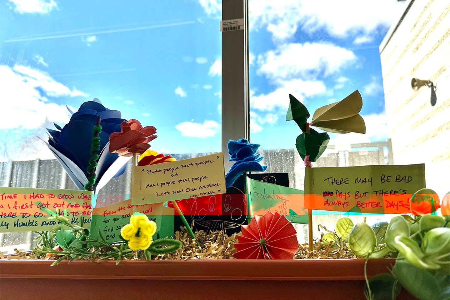 Paper flowers and notes with various phrases written on flags planted in a rectangular flower pot on a windowsill.