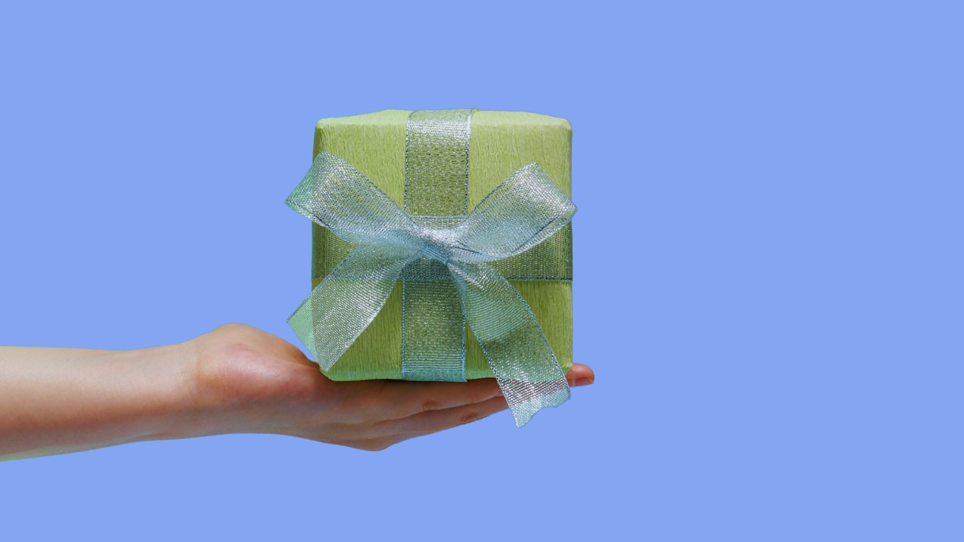 Hand holding out a present wrapped in green wrapping paper and ribbon against a blue background.