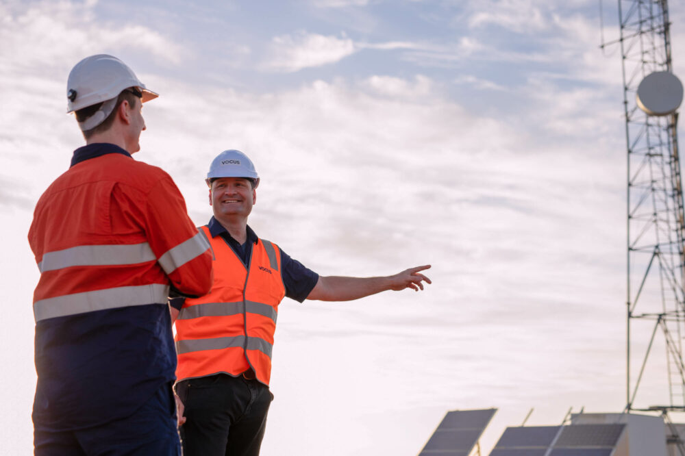 Photo of two Vocus workers in high vis and hard hats standing in front of a network tower at Minjar Gold site