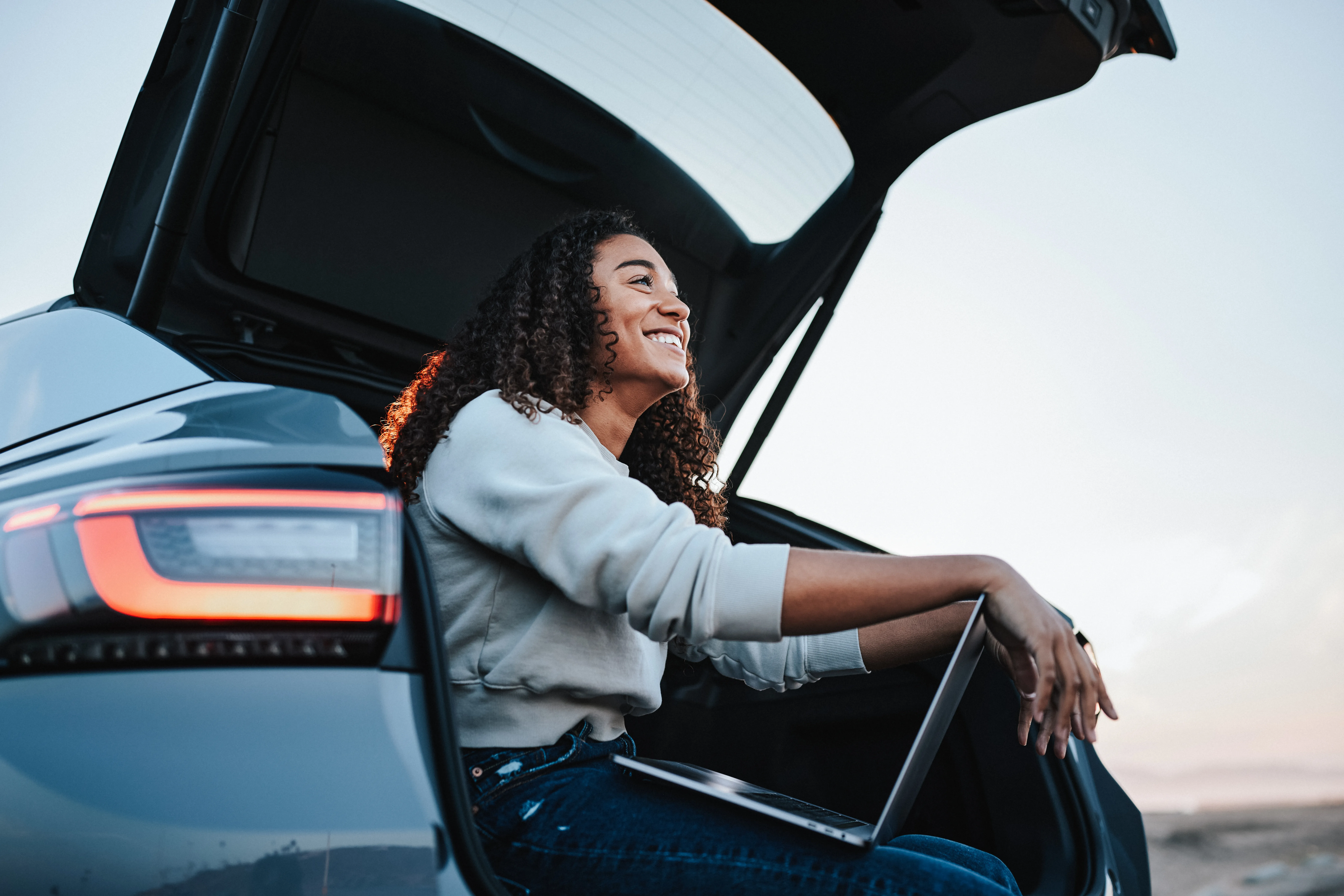 Happy woman with laptop sitting in car trunk, looking off into the distance. 