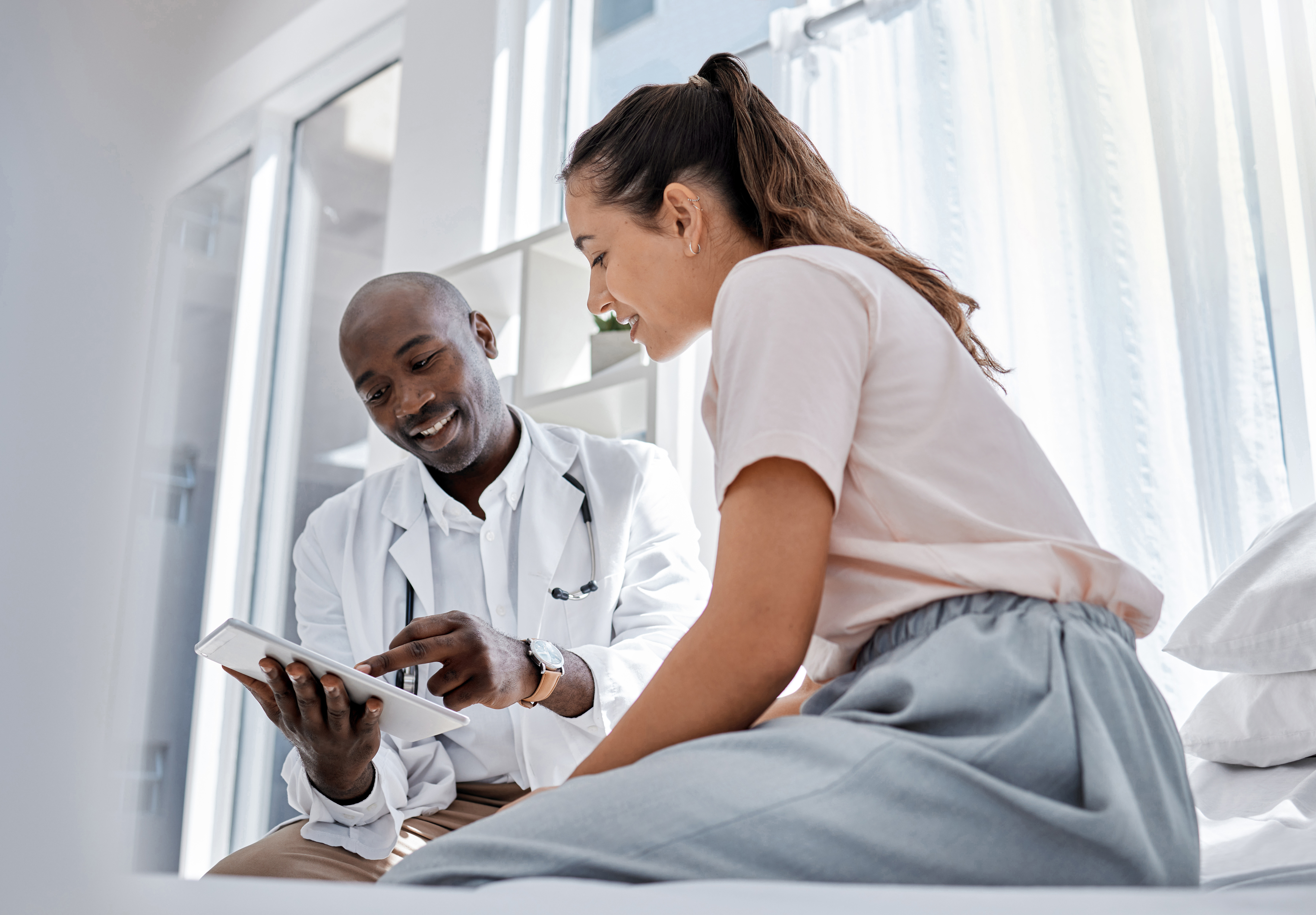 A male healthcare professional seated with a female patient in a bright clinical room, showing medical information on a tablet. 