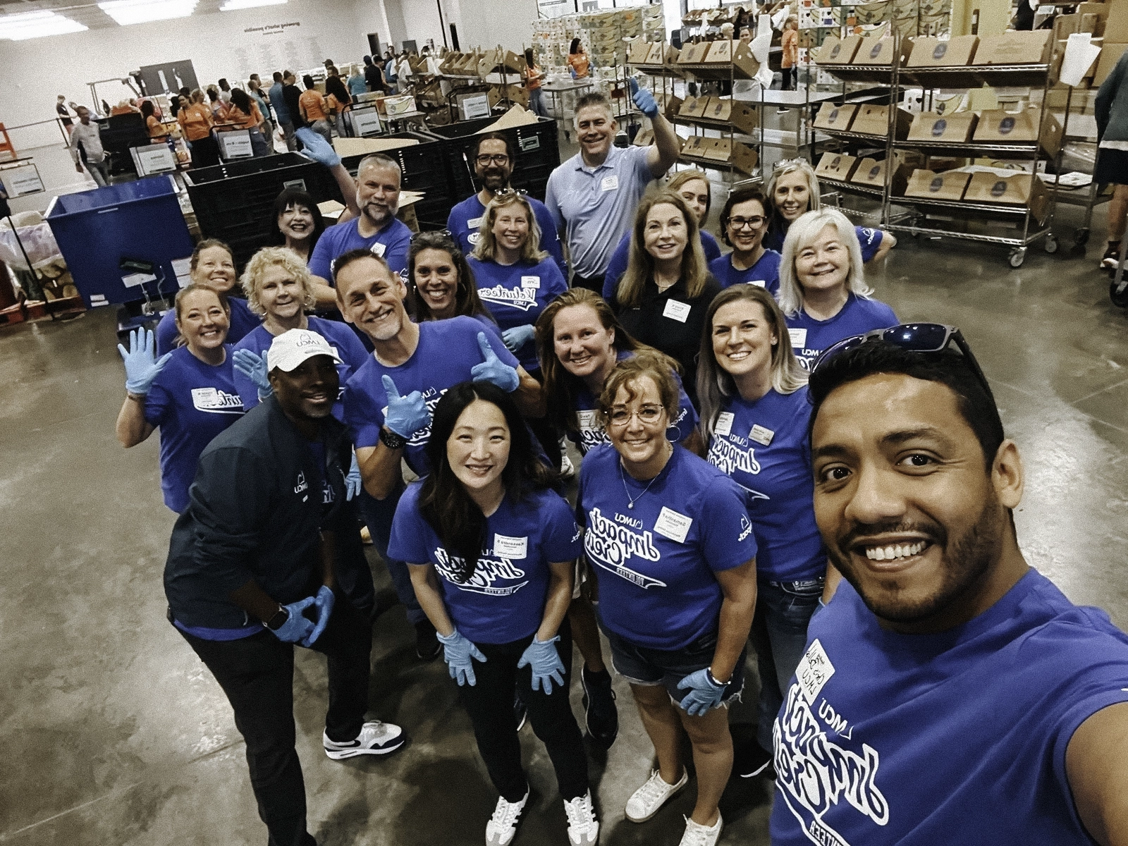 Group of volunteers in blue shirts gathered in a warehouse fulfillment center, posing for a photo among shelves of packaged goods and donation supplies.
